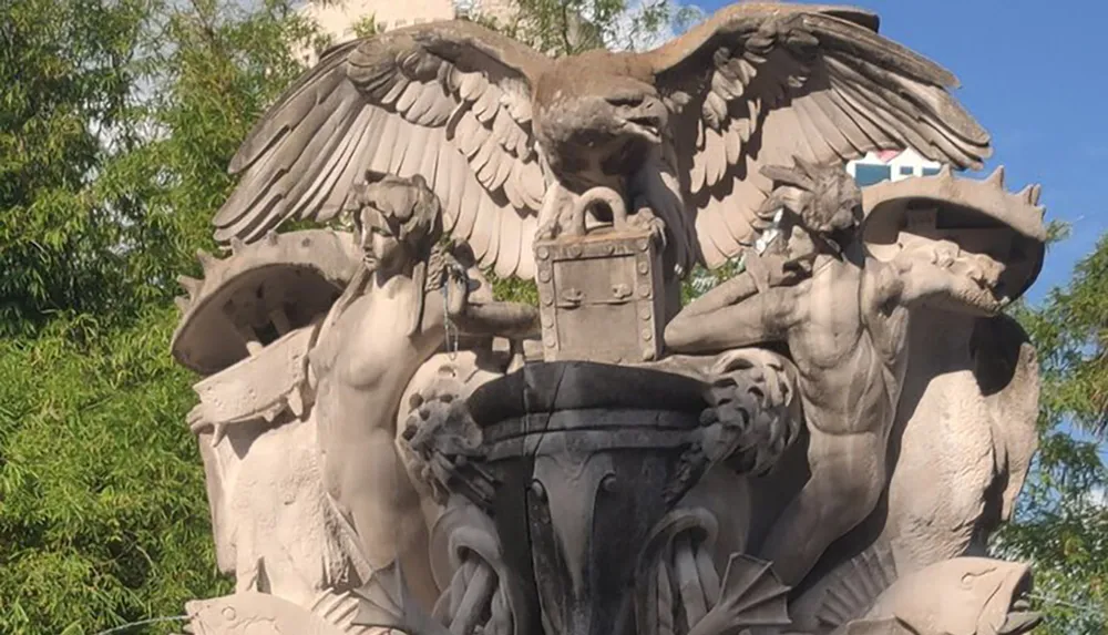 The image shows a sculpture featuring two figures and an eagle with outstretched wings centered above a shield and a decorative basin set against a background of green foliage