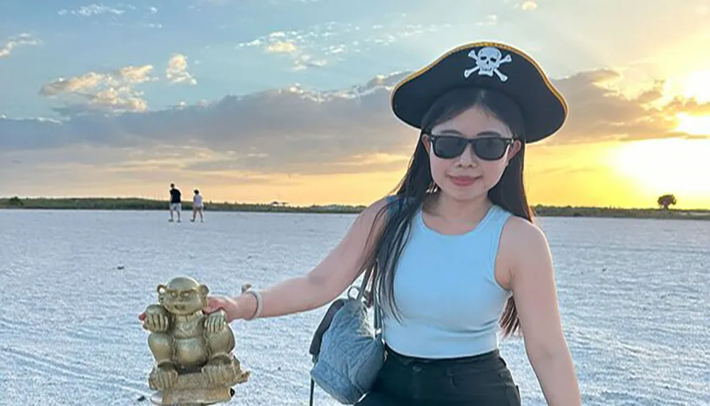 A person wearing a pirate hat and sunglasses is holding a small statue creating a playful optical illusion as if it were standing on the ground with two people in the background and a scenic sunset