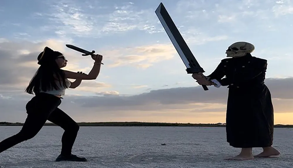 Two people one wearing a skull mask and holding a large foam sword and the other in a pirate hat with a small foam sword appear to be playfully dueling at sunset