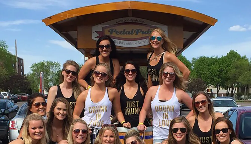 A group of smiling people wearing casual summer clothing stands in front of a Pedal Pub which is a multi-person bicycle designed for social outings