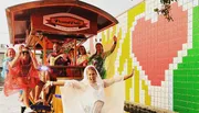 A group of cheerful people, some wearing ponchos, are enjoying a ride on a pedal pub, against a colorful mosaic-tiled wall background.