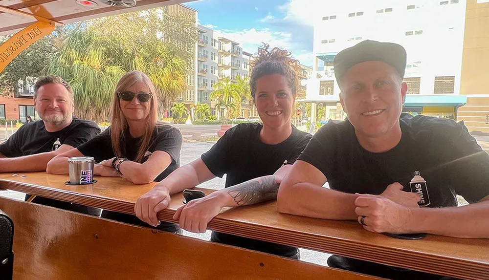 Four people are sitting at a wooden bar smiling and posing for the photo with a sunny urban backdrop