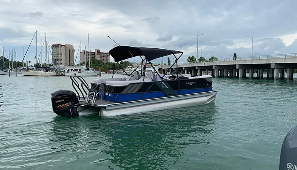 A pontoon boat with a black Bimini top is cruising near a bridge with several docked sailboats and buildings in the background