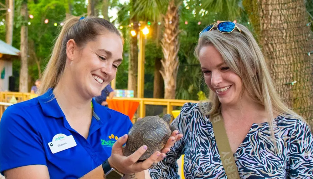 Two smiling women one in a blue zoo staff uniform share a moment while showing a small tortoise to the other