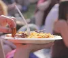 A person holds a plate of food with skewered meat and noodles in an outdoor setting with other people in the background