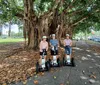 Three people are wearing helmets and standing on Segways under a large tree with hanging roots on a path littered with fallen leaves