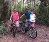 Three people are standing on a wooded trail with their bicycles smiling for the camera