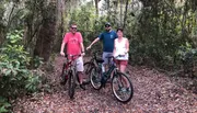 Three people are standing on a wooded trail with their bicycles, smiling for the camera.
