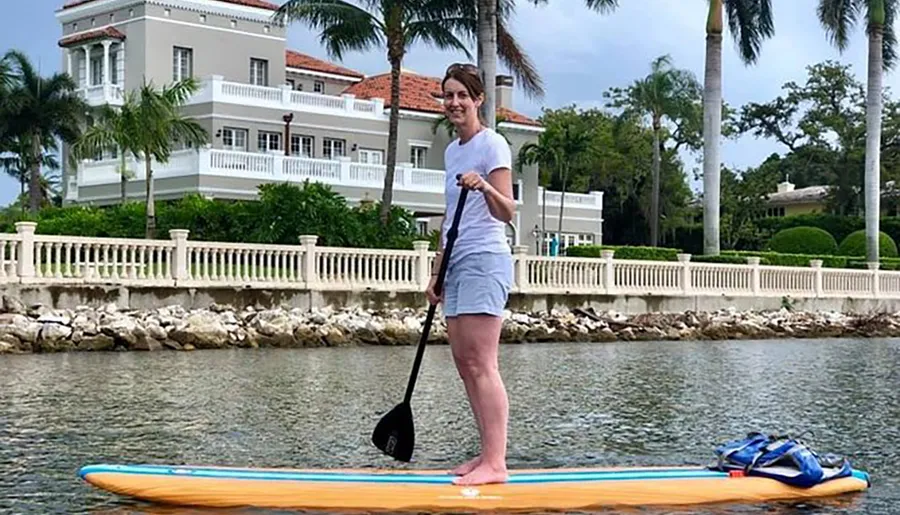 A person is standing on a paddleboard in calm water near a shoreline adorned with palm trees and a large house in the background.