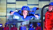 A person is enjoying an indoor skydiving experience with an instructor close by, while onlookers watch from behind a glass partition.