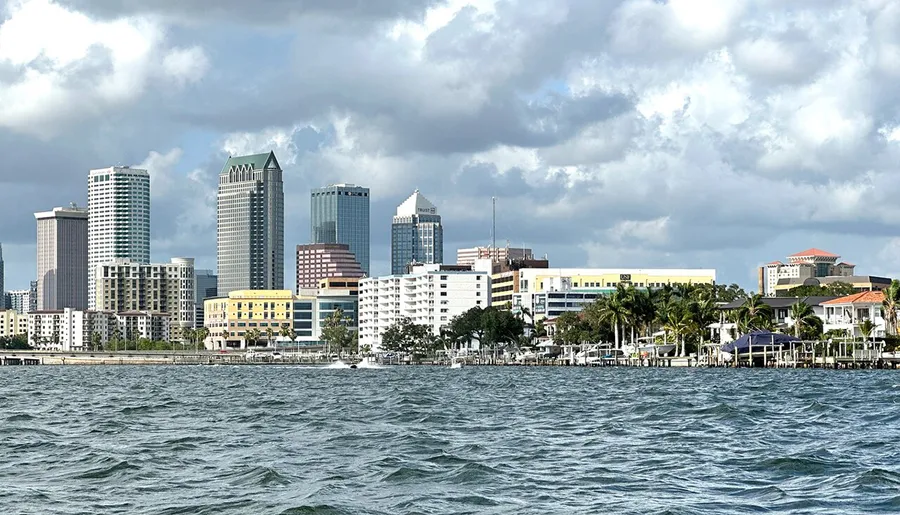 The image shows a picturesque view of a city skyline with various high-rise buildings, situated behind a body of water with gentle waves under a cloudy sky.
