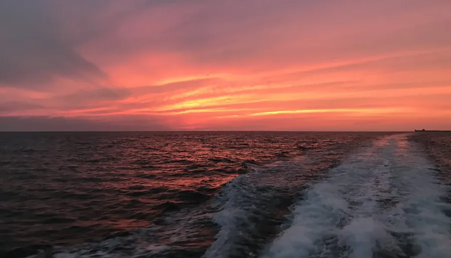 The image captures a striking sunset over the ocean with vivid orange and pink hues in the sky, reflected on the water's surface, and a wake from a boat visible in the foreground.
