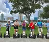 A group of people wearing helmets are lined up on Segways for a group photo with a backdrop of the city skyline and a marina