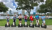 A group of people wearing helmets are lined up on Segways for a group photo with a backdrop of the city skyline and a marina.