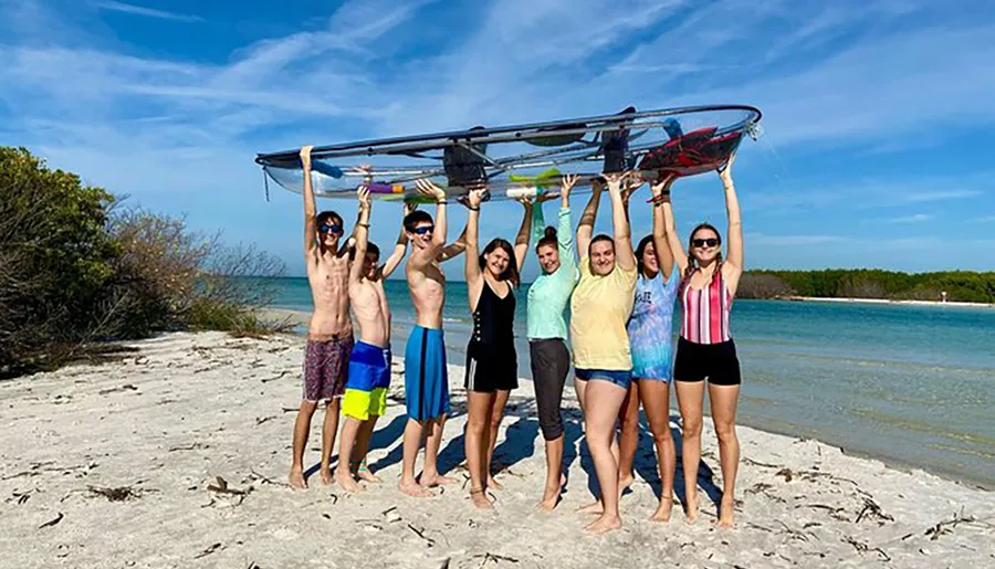 A group of people are holding a kayak above their heads on a sunny beach with a clear blue sky above them.