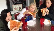 Three people are joyfully eating large slices of pizza at a table with drinks in red plastic cups.