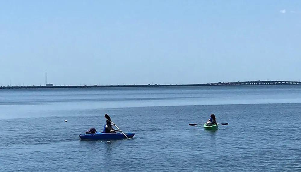 Two people are kayaking on calm waters with a bridge visible in the distant background