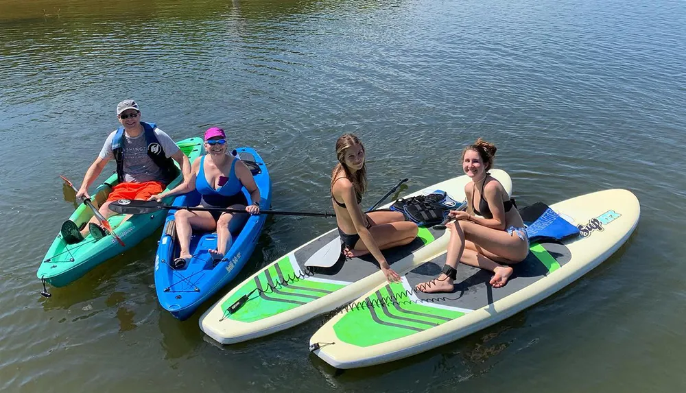 Four people are enjoying a sunny day on the water with kayaks and a paddleboard