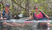 Two people are smiling and paddling in a clear kayak amongst mangrove trees.