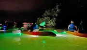A group of people are night kayaking with the water illuminated by underwater lights attached to their kayaks, creating a serene, glowing effect.