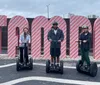 Three people are standing on Segways in front of a large striped sign that spells out TOYS with each person positioned inside one of the letters