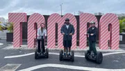Three people are standing on Segways in front of a large, striped sign that spells out 