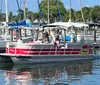 A group of people is enjoying a boat ride on a pontoon boat amidst a marina filled with various sailing boats on a sunny day