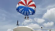 Two people are parasailing with a parachute decorated in a red, white, and blue pattern, likely enjoying a thrilling recreational activity on a sunny day.