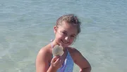 A smiling young girl is standing in shallow water at the beach, holding a sand dollar.