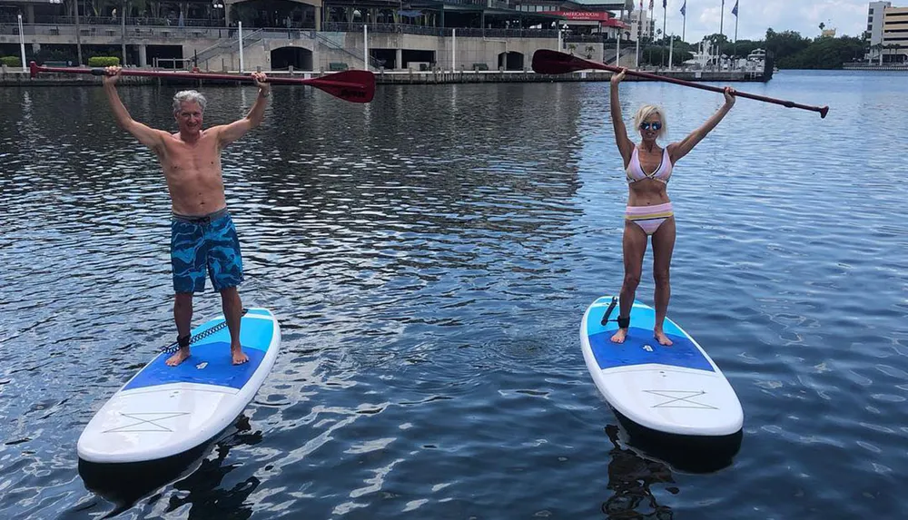 Two people are standing on paddleboards holding their paddles aloft on a calm water surface