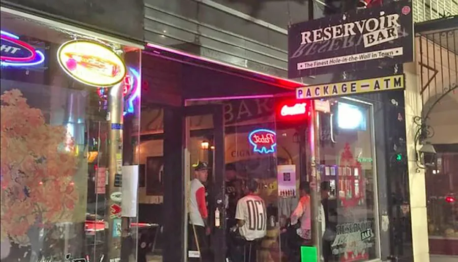 The image shows the exterior of the Reservoir Bar with neon signs and people standing by the entrance at night.