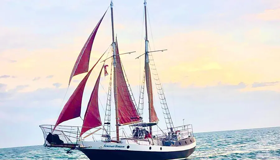 A sailboat with red sails is sailing on the sea against a soft pastel sky.