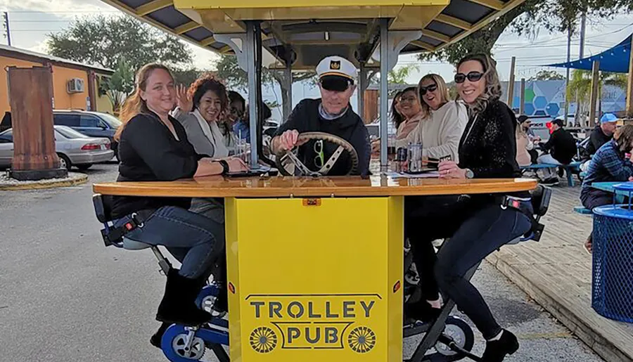 A group of people are enjoying themselves at a mobile bar called TROLLEY PUB, where a man steers while the customers sit around a table on bicycle-like seats.