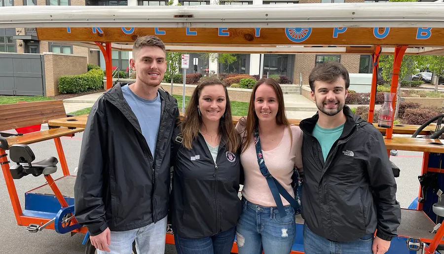 Four people are smiling in front of a vehicle named Trolley Pub that appears to be a pedal-powered mobile bar.