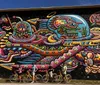 A group of cyclists are standing in front of a colorful intricate mural featuring psychedelic and vibrant imagery with the words Positive Creations and Peace Please