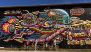 A group of cyclists are standing in front of a colorful, intricate mural featuring psychedelic and vibrant imagery with the words 
