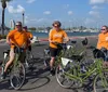 A group of people in orange shirts some with the University of Tennessee logo are riding or standing next to bicycles along a coastal area with palm trees and moored boats in the background