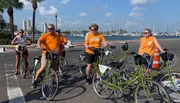 A group of people in orange shirts, some with the University of Tennessee logo, are riding or standing next to bicycles along a coastal area with palm trees and moored boats in the background.