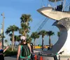 People are standing near a large artistic structure that resembles a ships propeller with palm trees and a clear blue sky in the background