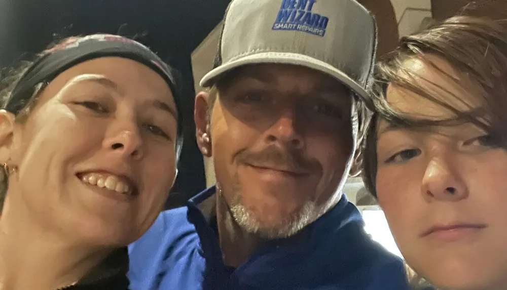 Three individuals are sharing a close-up selfie with one sporting a blue cap with a logo on it