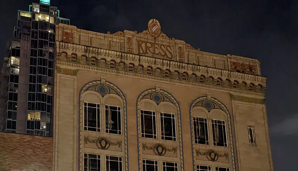 The image shows the ornate illuminated facade of the Kress building at night with a modern skyscraper in the background