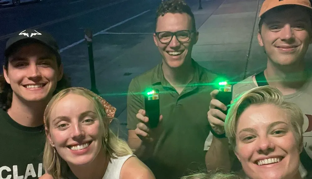 The image shows a group of five smiling people taking a group selfie at night with two of them holding devices that emit green lights