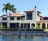 The image shows a large luxurious waterfront home with palm trees and a private dock under a bright blue sky
