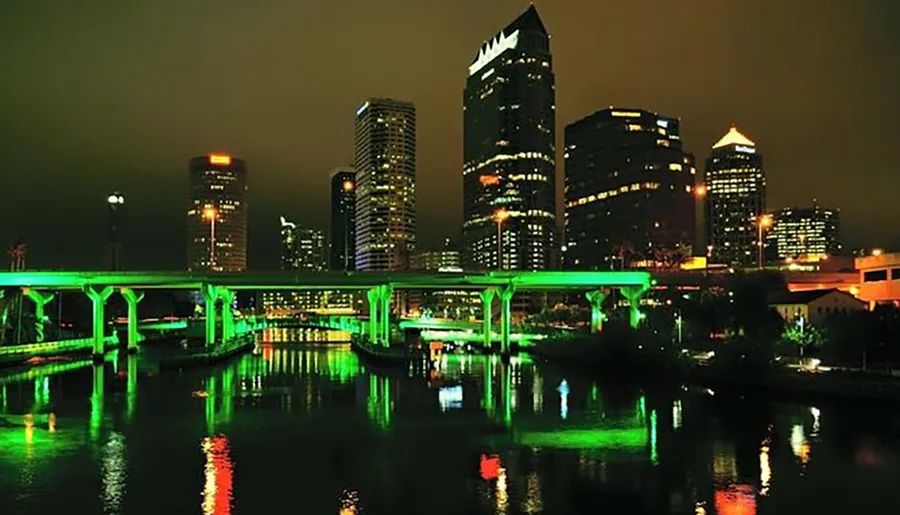 The image displays a nighttime view of a vibrant city skyline with illuminated buildings reflecting off the water, complemented by a brightly lit green bridge that spans across the river.