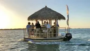 A group of people enjoys a gathering on a floating tiki bar boat at sunset.