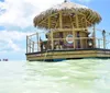 The image depicts a thatch-roofed floating tiki bar surrounded by clear turquoise waters under a blue sky with people enjoying the unique setting on the water