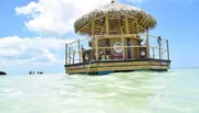 The image depicts a thatch-roofed floating tiki bar surrounded by clear turquoise waters under a blue sky, with people enjoying the unique setting on the water.