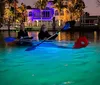 Two people are kayaking at dusk on water illuminated by the glow from their clear kayak with an illuminated house in the background