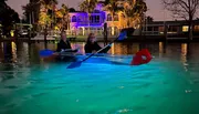 Two people are kayaking at dusk on water illuminated by the glow from their clear kayak, with an illuminated house in the background.