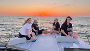 A group of individuals is enjoying a sunset on a boat, with calm seas and a colorful sky in the background.